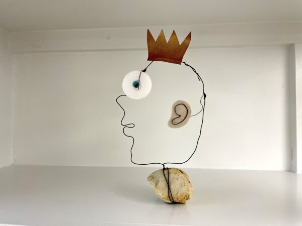 A minimalist wire sculpture of a human head in profile, featuring a brown paper crown, a white circular eye with a blue center, a beige ear, and a rock base, displayed against a plain white background.