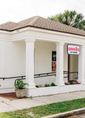 A small white building with columns and ramp access, displaying a sign that reads amelia art studio on the front and side. Lush greenery and potted plants decorate the entrance.