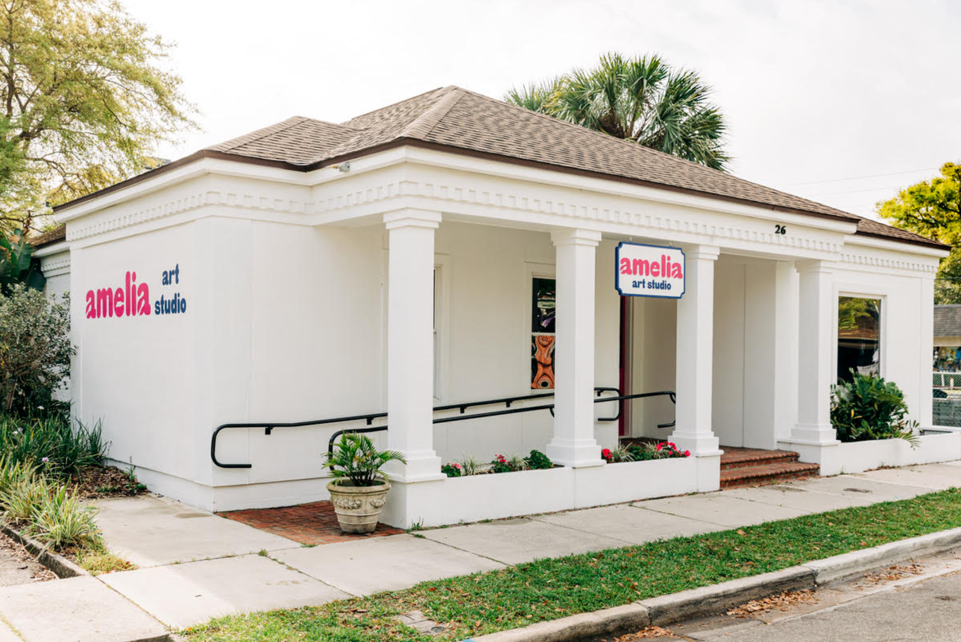 A small white building with columns and ramp access, displaying a sign that reads amelia art studio on the front and side. Lush greenery and potted plants decorate the entrance.