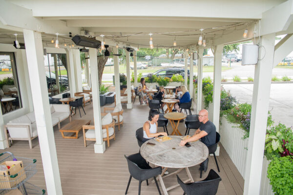 People sit at round tables on a covered patio with string lights, eating and talking. There are plants, modern outdoor furniture, and cars parked outside in the background.
