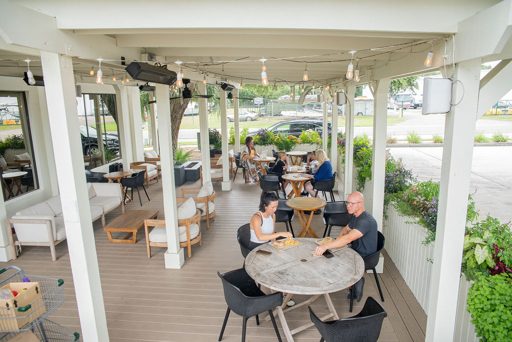 People sit at round tables on a covered patio with string lights, eating and talking. There are plants, modern outdoor furniture, and cars parked outside in the background.
