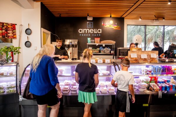 Three children and an adult stand at a café counter, looking at food in a display case. A staff member is behind the counter. The café is brightly lit with snacks and drinks visible around the space.
