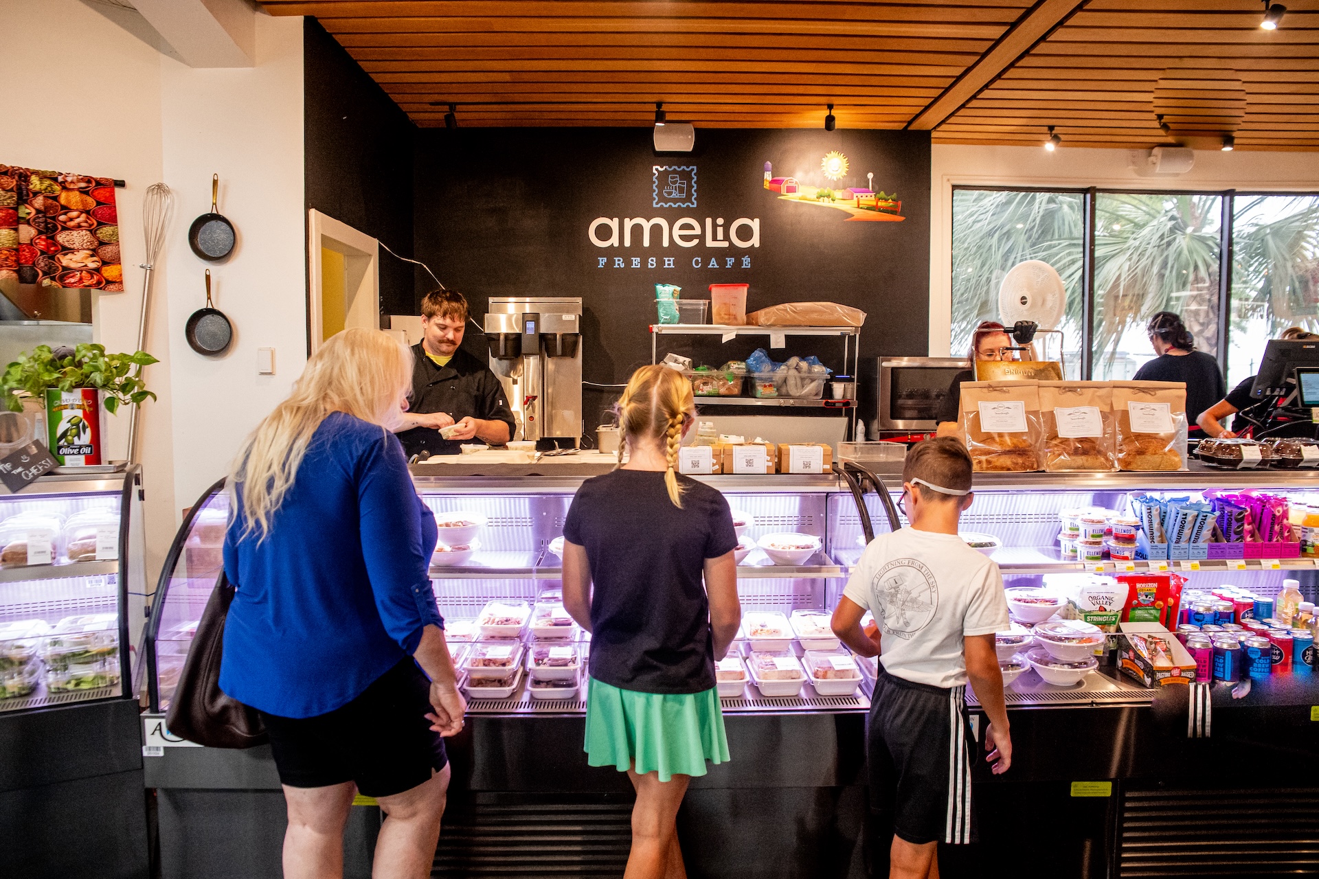 Three children and an adult stand at a café counter, looking at food in a display case. A staff member is behind the counter. The café is brightly lit with snacks and drinks visible around the space.