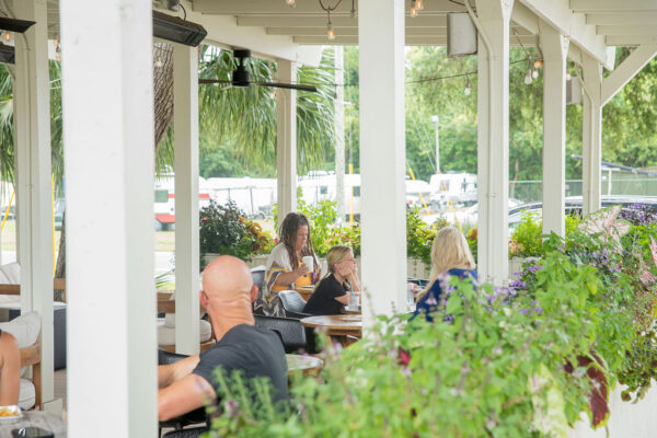 People sit at outdoor tables under a covered patio surrounded by green plants and flowers, enjoying food and drinks on a sunny day. Cars and trees are visible in the background through the open sides.