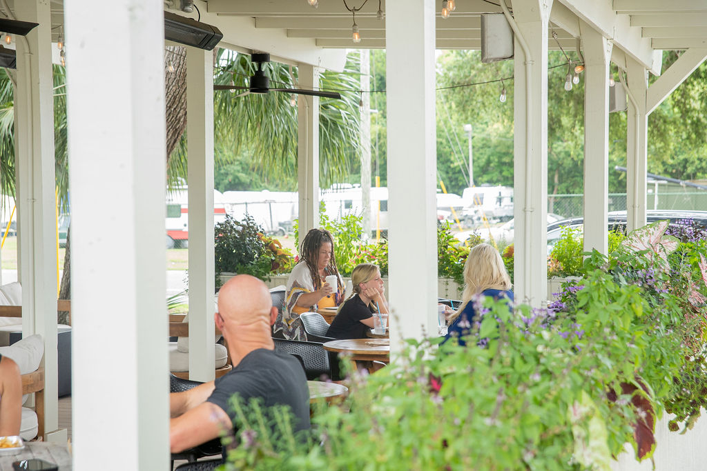 People sit at outdoor tables under a covered patio surrounded by green plants and flowers, enjoying food and drinks on a sunny day. Cars and trees are visible in the background through the open sides.