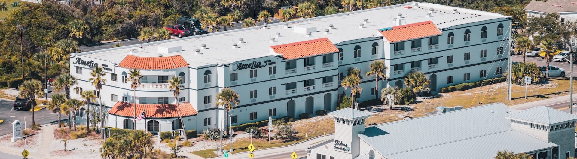 Aerial view of a white, three-story hotel with red-tiled roof accents, palm trees, and a busy parking lot, situated near a main road and surrounded by greenery and other buildings.