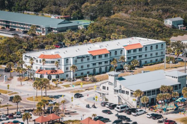 Aerial view of a white, three-story hotel with red-tiled roof accents, palm trees, and a busy parking lot, situated near a main road and surrounded by greenery and other buildings.