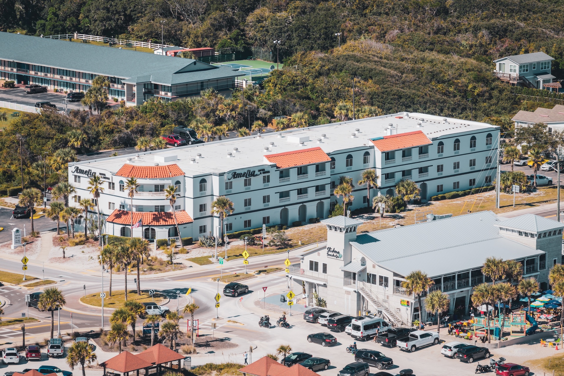Aerial view of a white, three-story hotel with red-tiled roof accents, palm trees, and a busy parking lot, situated near a main road and surrounded by greenery and other buildings.