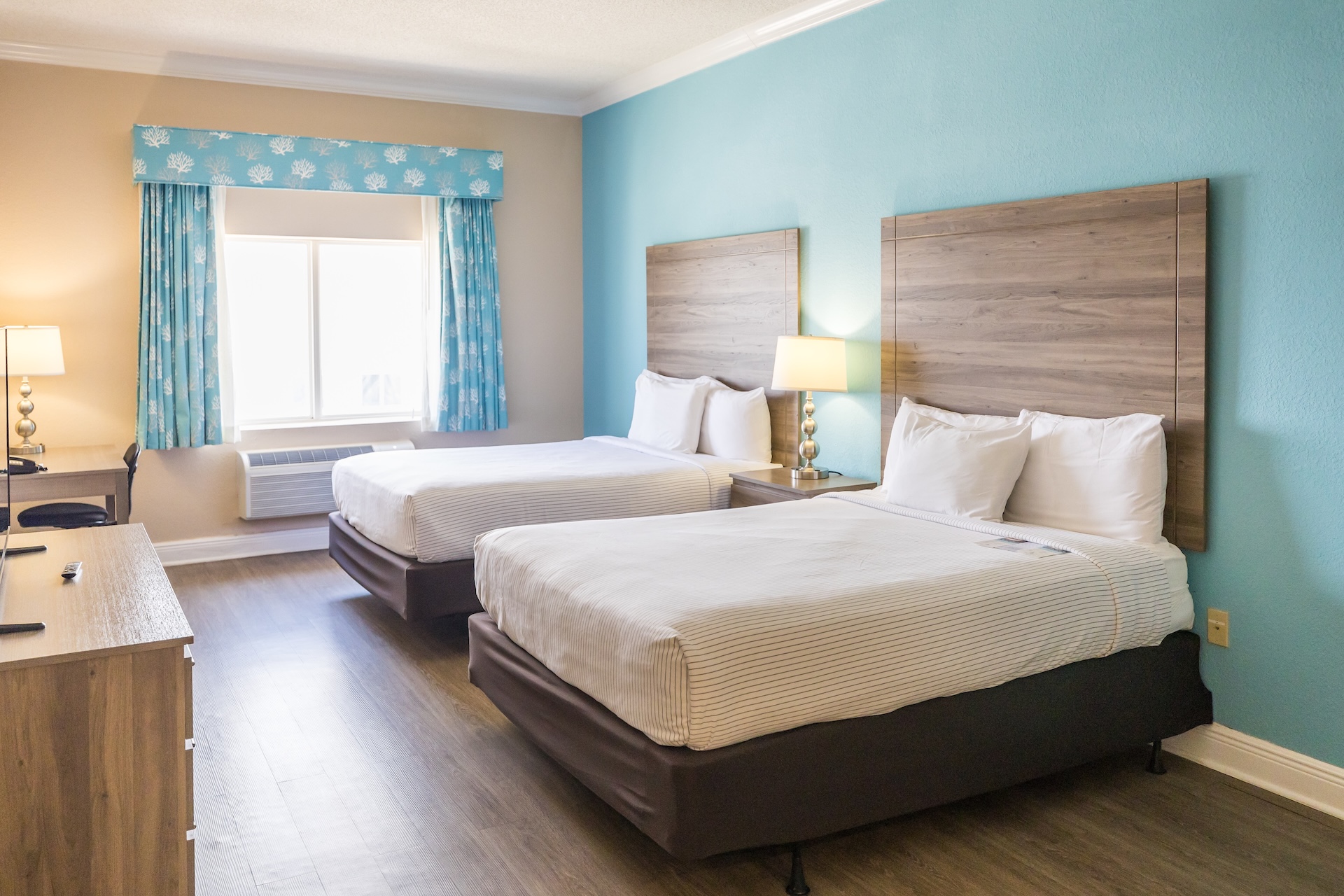 A hotel room with two neatly made double beds, wooden headboards, a nightstand with a lamp, a desk, a TV, and a window with blue-patterned curtains. The walls are beige and light blue, and the floor is wood.