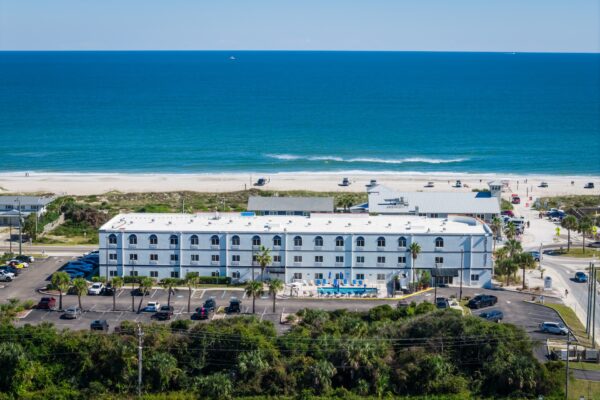 Aerial view of a white, two-story beachfront hotel with a swimming pool in front, palm trees, parked cars, sandy beach, and the blue ocean in the background under a clear sky.