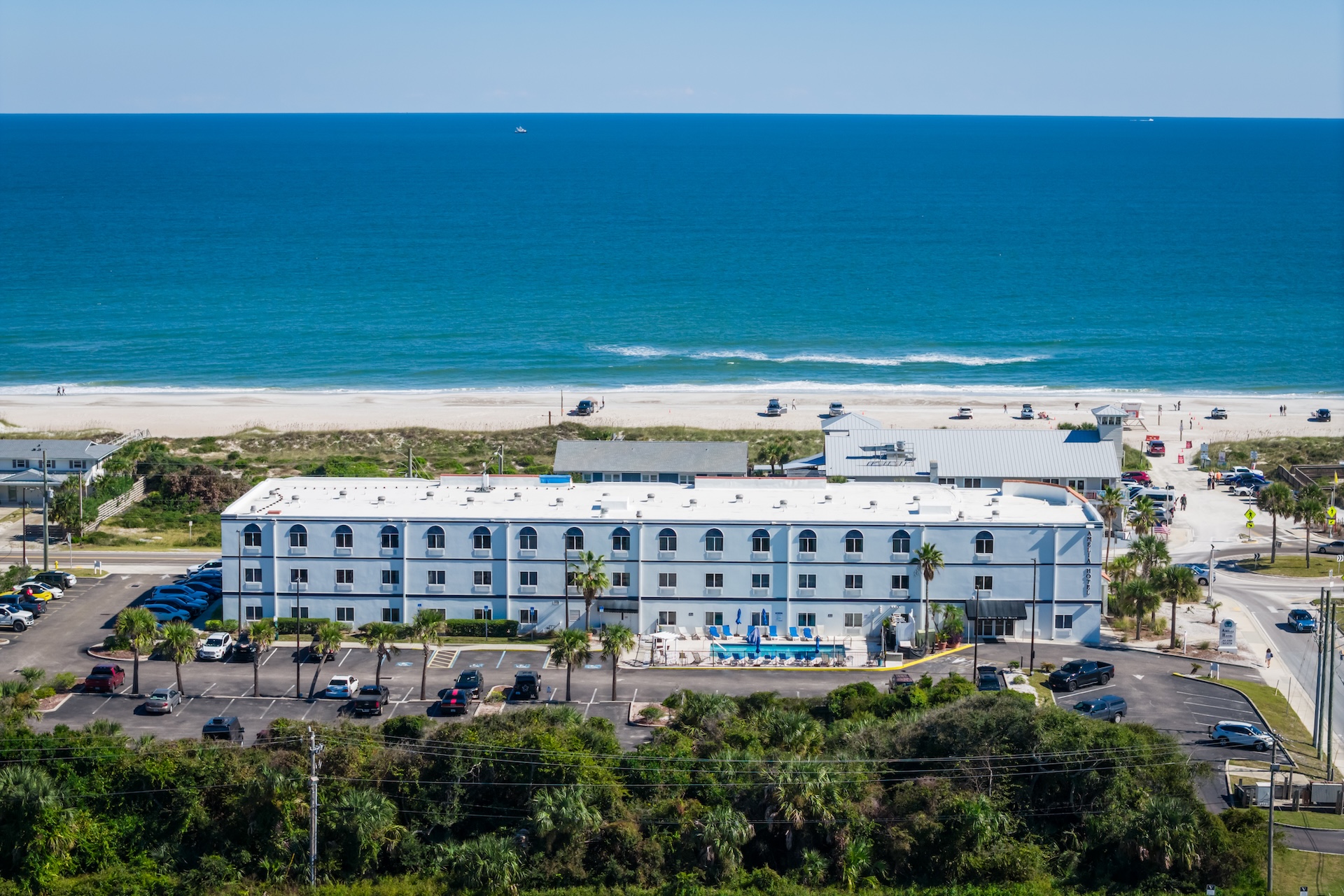 Aerial view of a white, two-story beachfront hotel with a swimming pool in front, palm trees, parked cars, sandy beach, and the blue ocean in the background under a clear sky.