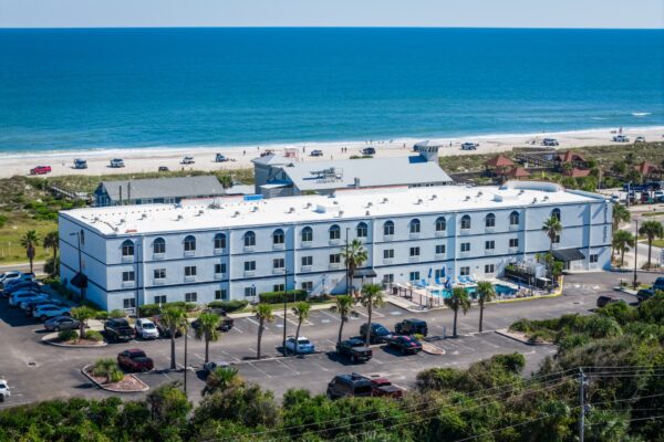 Aerial view of a white three-story hotel with a parking lot, palm trees, and swimming pool, located near a sandy beach and blue ocean under a clear sky.