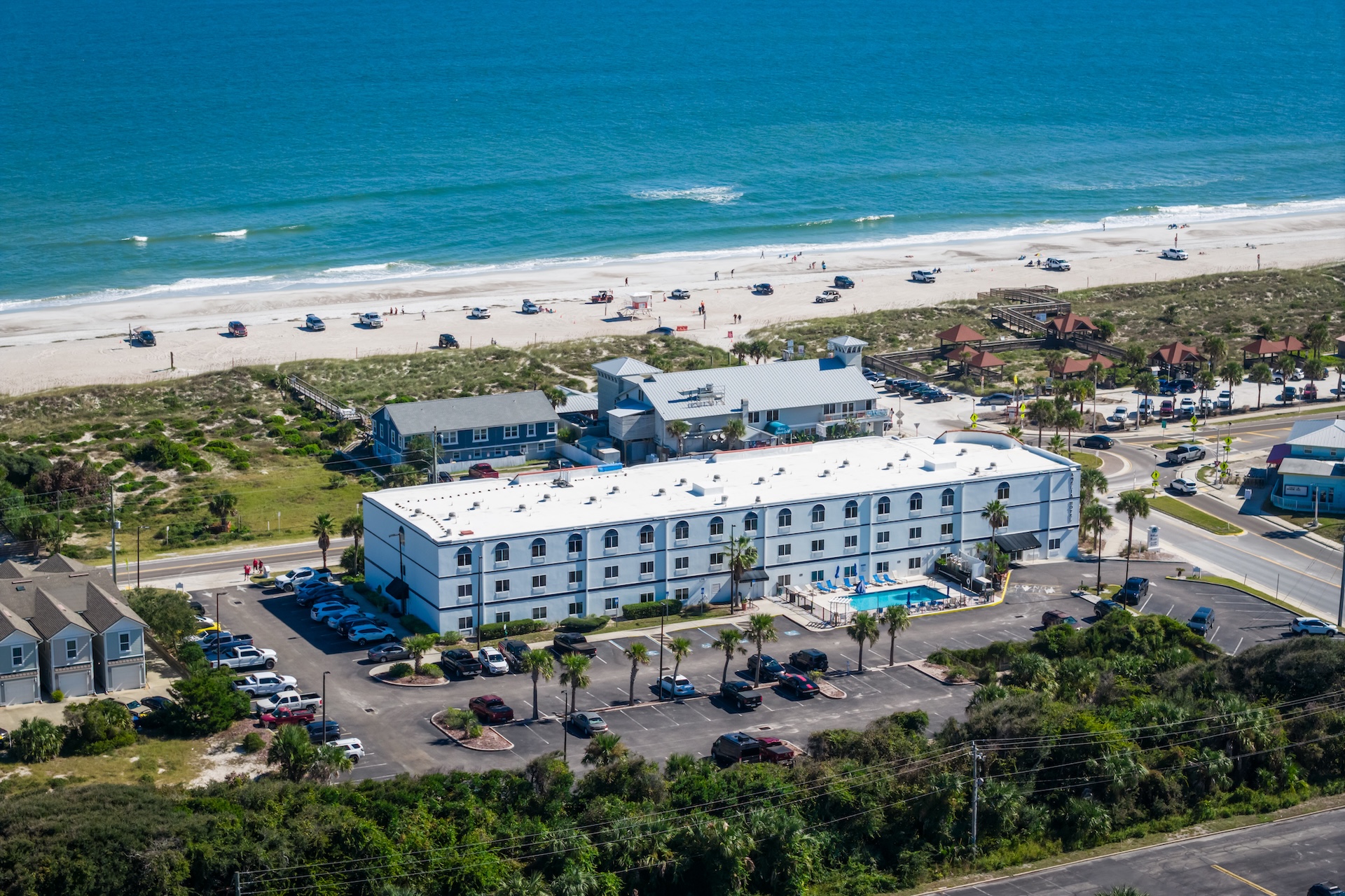 Aerial view of a beachfront hotel with a white roof, surrounded by parked cars and palm trees, facing a sandy beach and the ocean; people are visible on the beach and near the water.