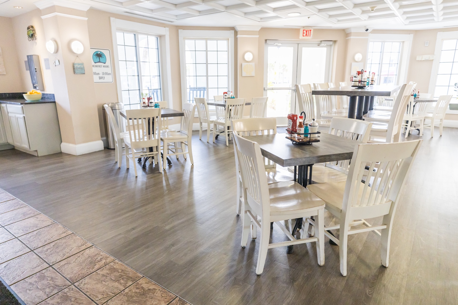 Bright, airy dining area with white wooden tables and chairs, large windows letting in natural light, neutral walls, condiment caddies on tables, and a tiled floor section near the entrance.