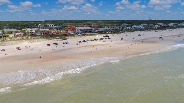 Aerial view of a sandy beach with gentle waves, scattered people, and parked cars. Buildings and greenery line the background beneath a partly cloudy blue sky.