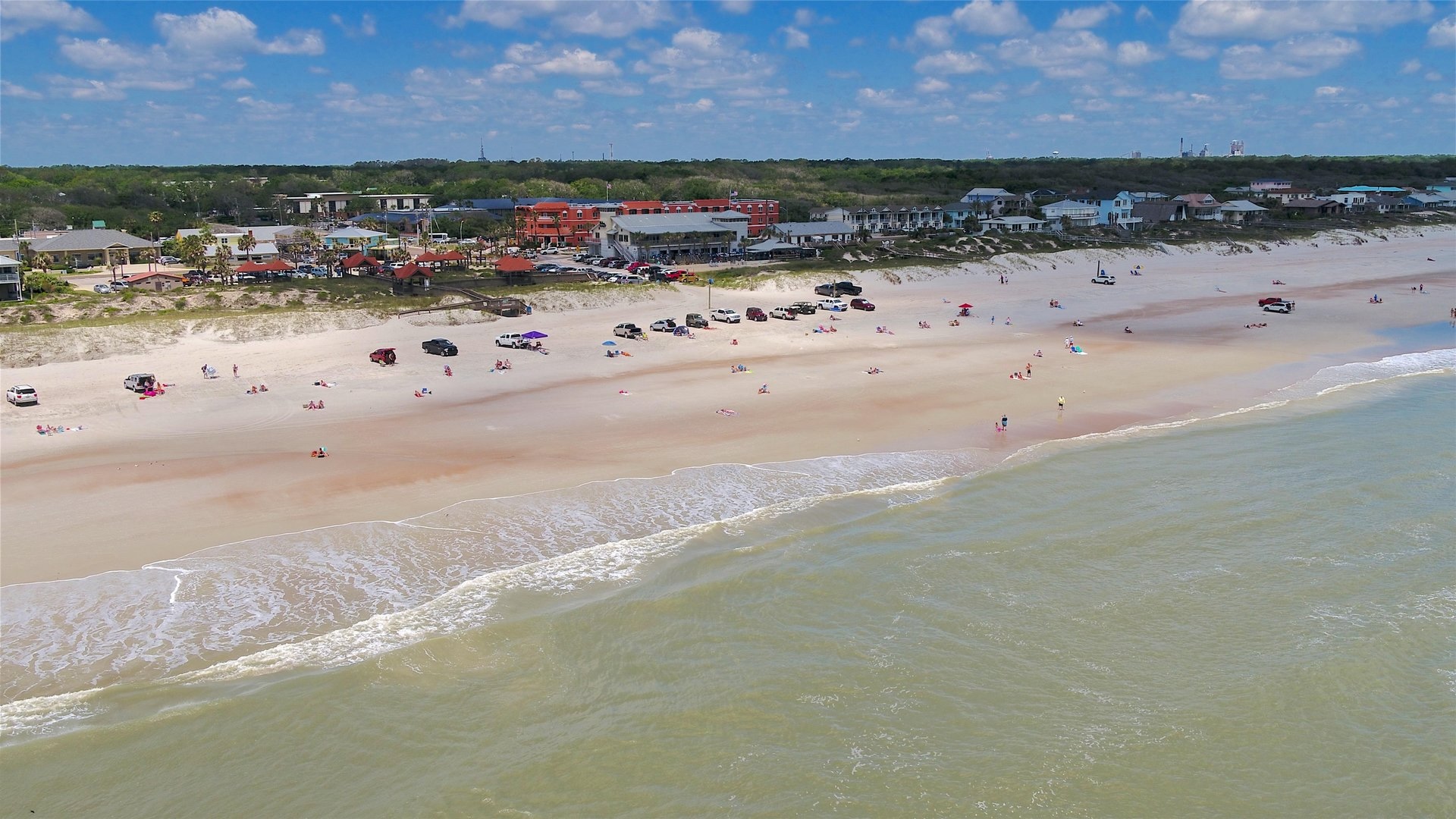 Aerial view of a sandy beach with gentle waves, scattered people, and parked cars. Buildings and greenery line the background beneath a partly cloudy blue sky.