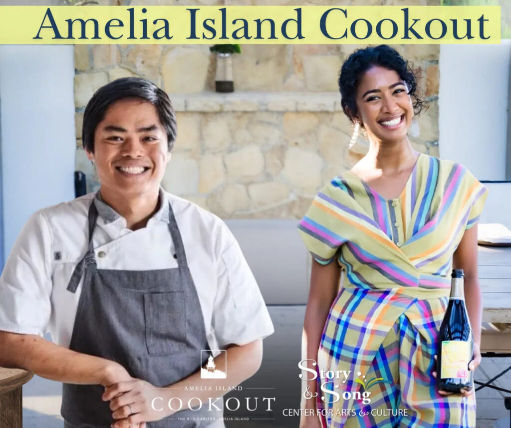 Two smiling people stand side by side at the Amelia Island Cookout event; the man on the left wears a chefs coat and apron, while the woman on the right holds a wine bottle and wears a colorful striped dress.