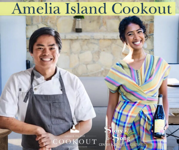 Two smiling people stand side by side at the Amelia Island Cookout event; the man on the left wears a chefs coat and apron, while the woman on the right holds a wine bottle and wears a colorful striped dress.