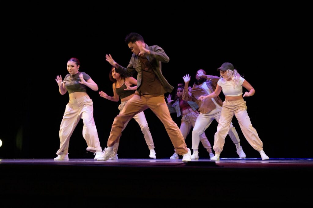 Six dancers perform a hip-hop routine on a dark stage, all wearing casual outfits and sneakers. The group is mid-motion, expressing intense energy and focus while synchronized in dynamic poses.