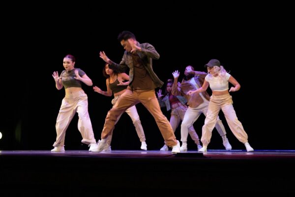 Six dancers perform a hip-hop routine on a dark stage, all wearing casual outfits and sneakers. The group is mid-motion, expressing intense energy and focus while synchronized in dynamic poses.