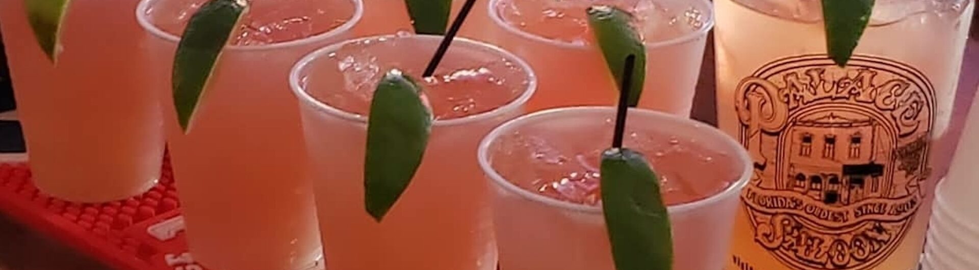 Eight plastic cups filled with a light orange drink, each garnished with a lime wedge and a black straw, are lined up on a bar mat. A tall glass with a logo and a straw is beside them.