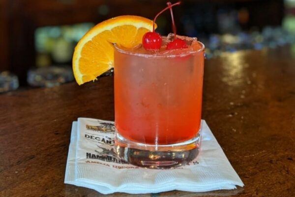 A red cocktail in a glass garnished with two maraschino cherries and an orange slice, placed on a white napkin on a wooden bar counter.