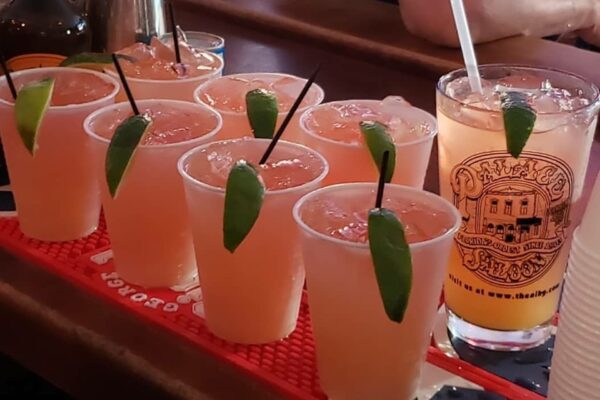 Eight plastic cups filled with a light orange drink, each garnished with a lime wedge and a black straw, are lined up on a bar mat. A tall glass with a logo and a straw is beside them.