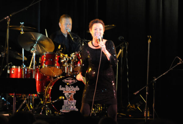 A woman in a black sequined dress sings into a microphone on stage, with a drummer playing a red drum set in the background. Stage lights shine on them, and a Jazz sign is visible on the drum kit.