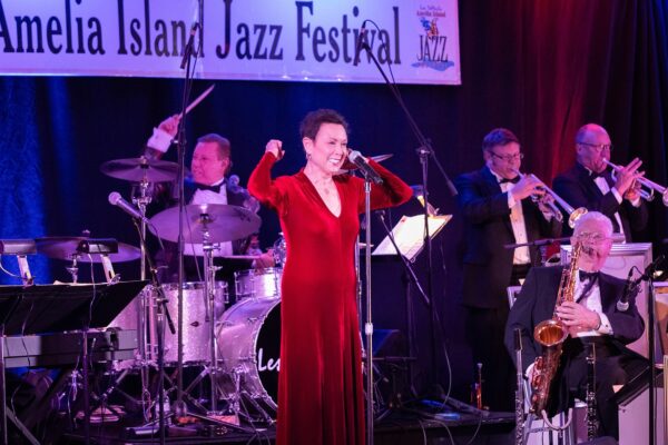 A woman in a red dress sings on stage with a jazz band at the Amelia Island Jazz Festival. Musicians play drums, trumpet, saxophone, and piano behind her, with festival signage visible above them.