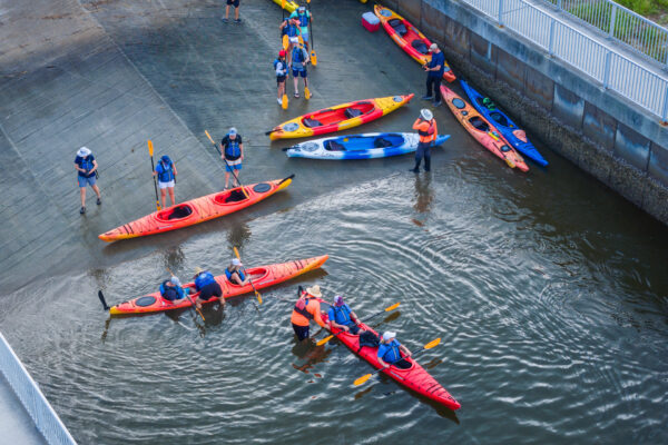 A group of people wearing life jackets prepare to launch colorful kayaks from a concrete ramp into the water. Some are standing on the ramp while others paddle in the river near the edge.