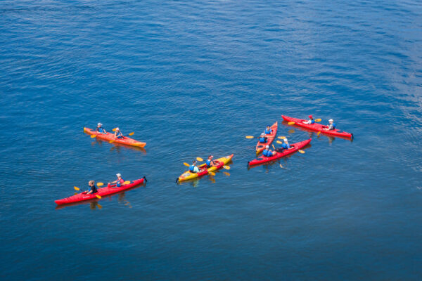 A group of people in red and yellow kayaks paddle together on calm blue water, wearing life jackets and hats.
