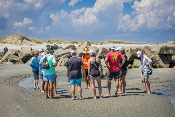 A group of people stands barefoot in a circle on a sandy beach near rocks and shallow water, listening to a person in an orange vest under a sunny, partly cloudy sky.