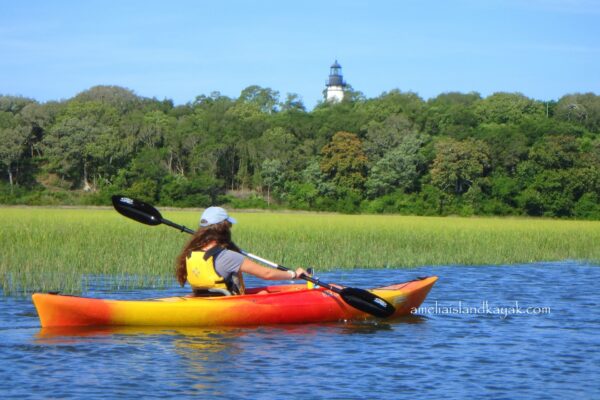 A person in a red and yellow kayak paddles on calm water, surrounded by green marsh grass, with a lighthouse visible in the background against a backdrop of trees.