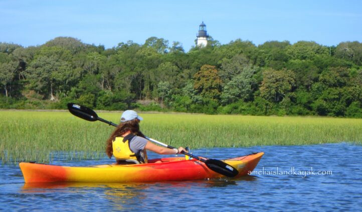 A person in a red and yellow kayak paddles on calm water, surrounded by green marsh grass, with a lighthouse visible in the background against a backdrop of trees.