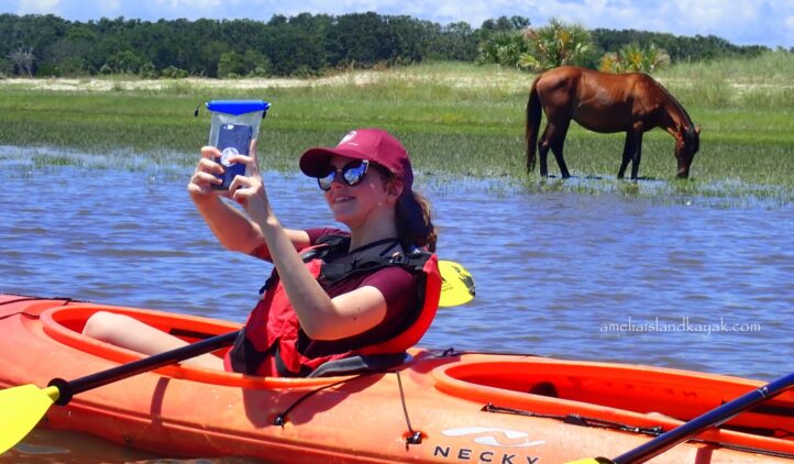 A woman in a red kayak takes a selfie with her phone, smiling and wearing sunglasses and a cap. A brown horse grazes on grass by the water in the background under a blue sky.