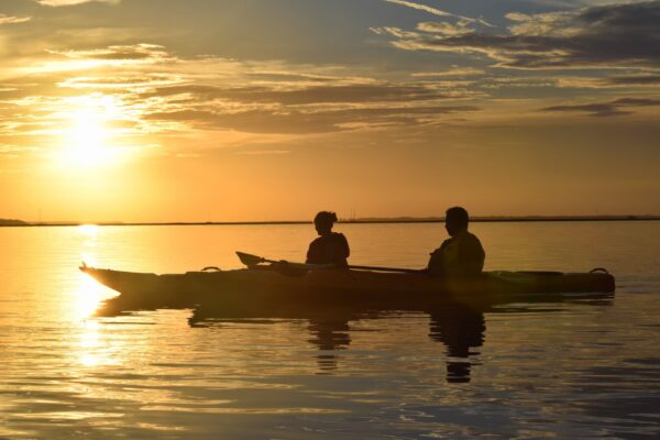 Two people in a kayak paddle on calm water at sunset, silhouetted against a golden sky with scattered clouds. The sunlight reflects off the water, creating a peaceful and serene scene.