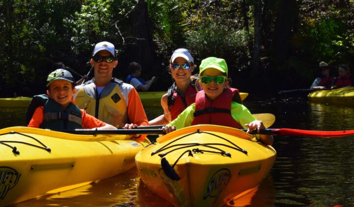 A smiling family of four in bright shirts and hats sits in two yellow kayaks on a sunlit, wooded river, enjoying a day of paddling together.