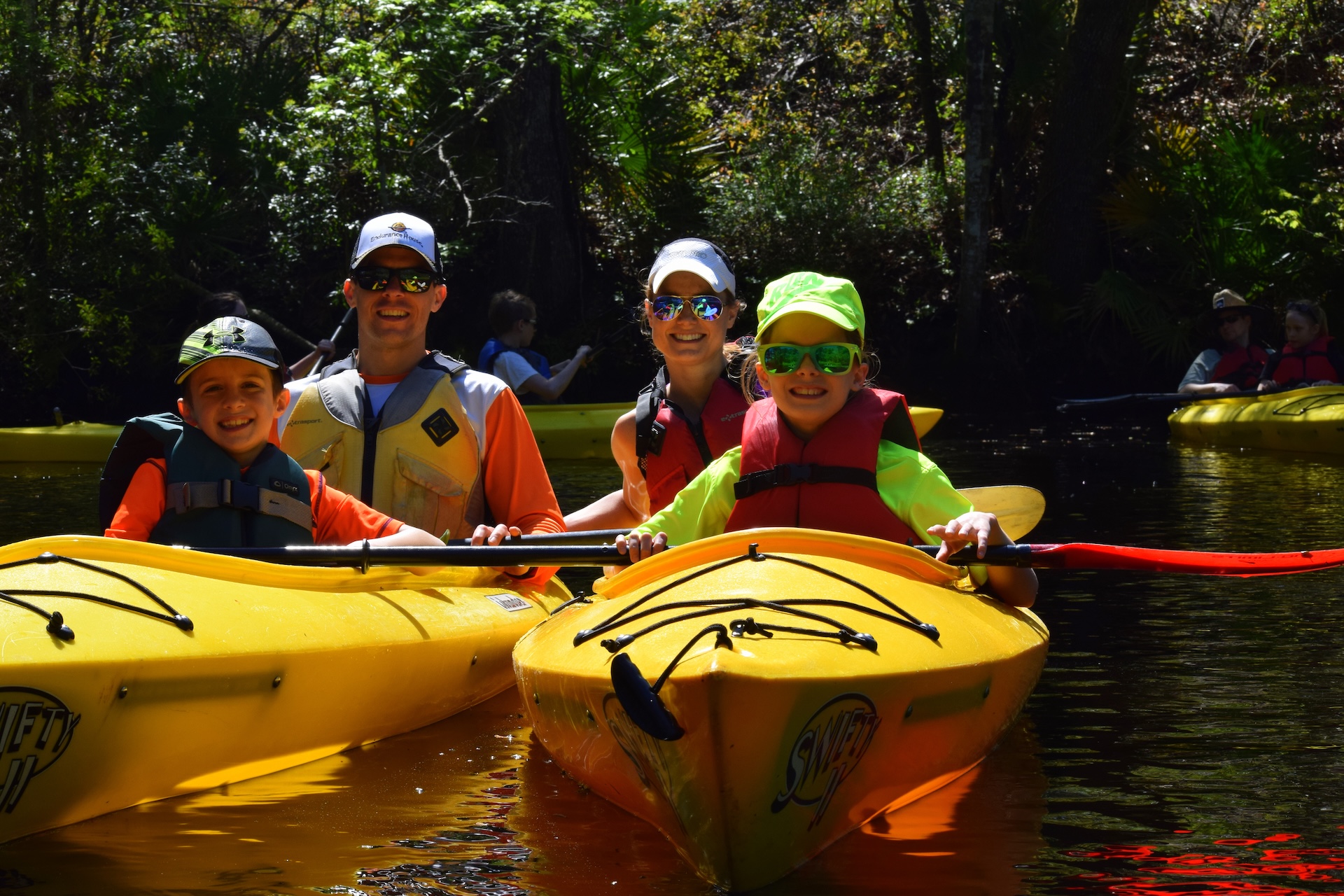 A smiling family of four in bright shirts and hats sits in two yellow kayaks on a sunlit, wooded river, enjoying a day of paddling together.
