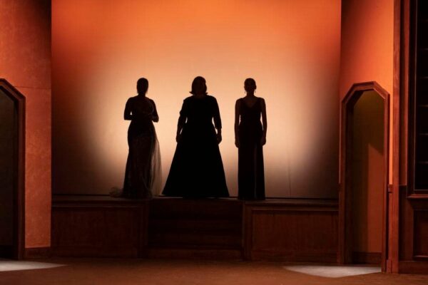 Three women in formal gowns stand silhouetted against a warm, orange-lit background on a stage, framed by dark wooden set pieces at the sides.