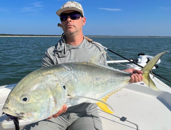 A person wearing sunglasses and a cap holds a large fish while sitting on a boat in open water, with land visible in the distance under a clear blue sky.