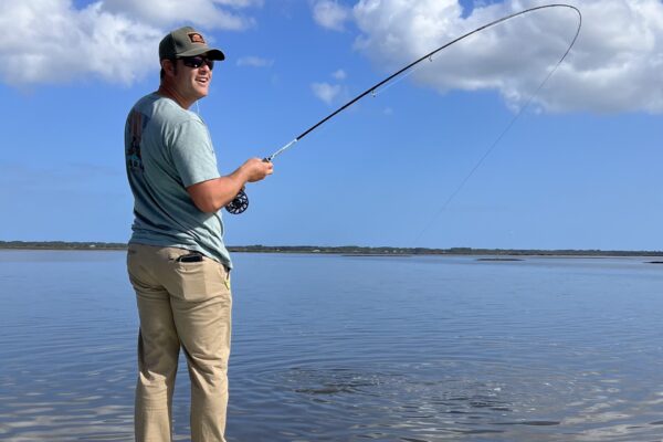 A man wearing sunglasses, a cap, and a light blue shirt is standing in shallow water while fly fishing. He is smiling and holding a fishing rod, with blue sky and clouds in the background.