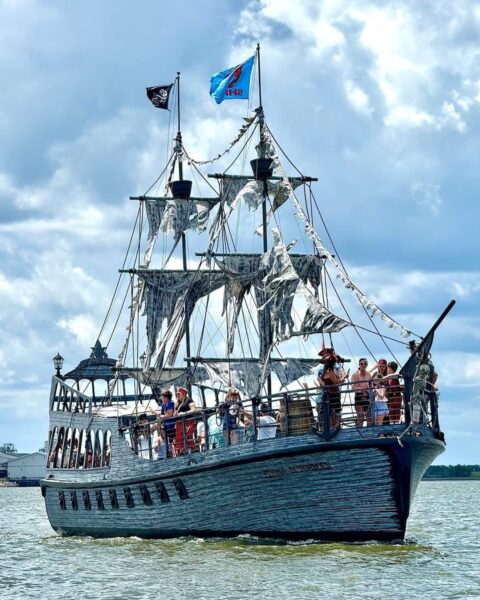 A large pirate-themed ship with tattered sails carries a group of people on the water. The ship has two flags, including a pirate flag, and appears to be for tours or entertainment. The sky is partly cloudy.