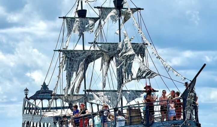 A large pirate-themed ship with tattered sails carries a group of people on the water. The ship has two flags, including a pirate flag, and appears to be for tours or entertainment. The sky is partly cloudy.