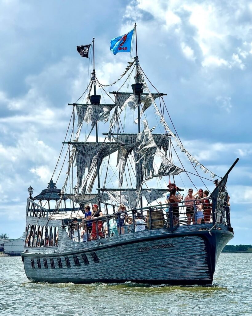 A large pirate-themed ship with tattered sails carries a group of people on the water. The ship has two flags, including a pirate flag, and appears to be for tours or entertainment. The sky is partly cloudy.