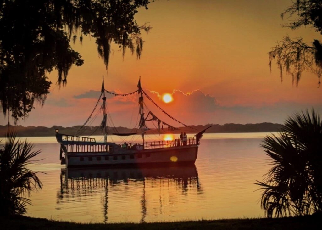 A silhouetted pirate ship floats on calm water at sunset, surrounded by trees and Spanish moss, with the sky glowing orange and pink over the horizon.