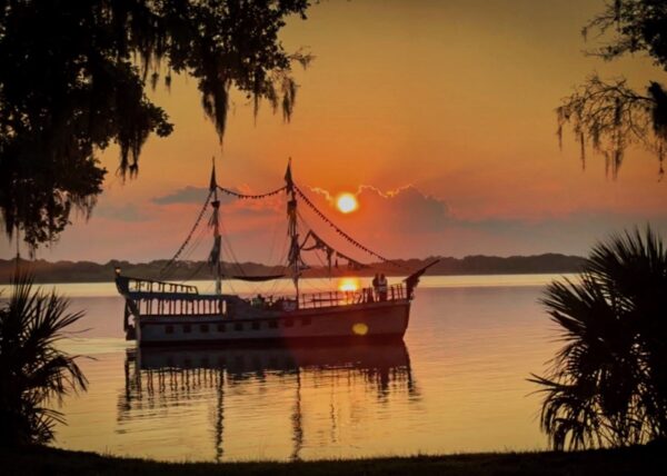 A silhouetted pirate ship floats on calm water at sunset, surrounded by trees and Spanish moss, with the sky glowing orange and pink over the horizon.