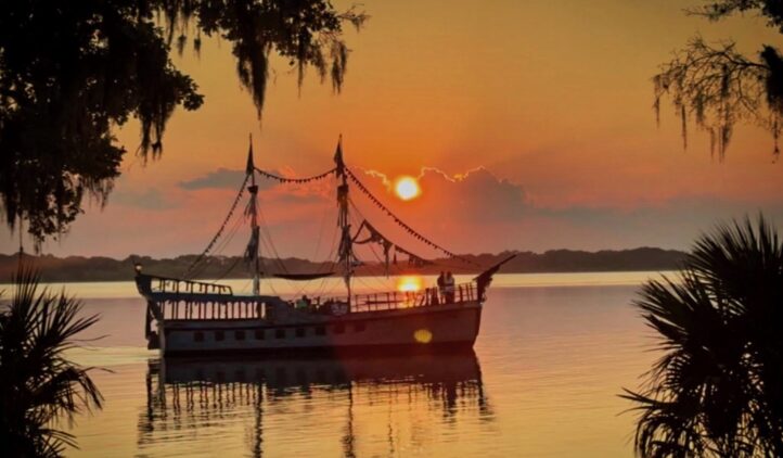 A silhouetted pirate ship floats on calm water at sunset, surrounded by trees and Spanish moss, with the sky glowing orange and pink over the horizon.