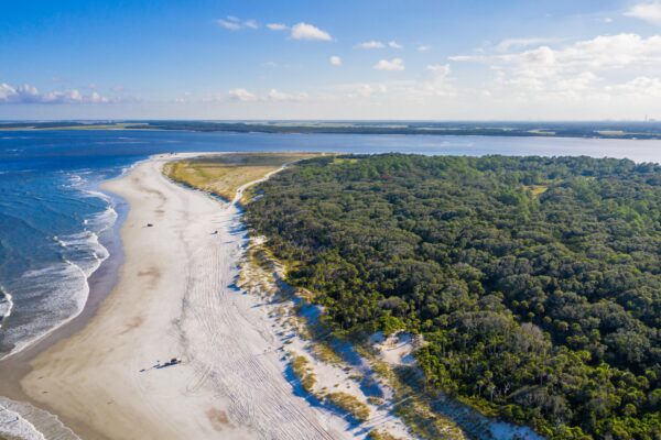 Aerial view of a sandy beach curving along the shoreline, bordered by dense green forest under a blue sky with scattered clouds. The ocean waves gently meet the shore.