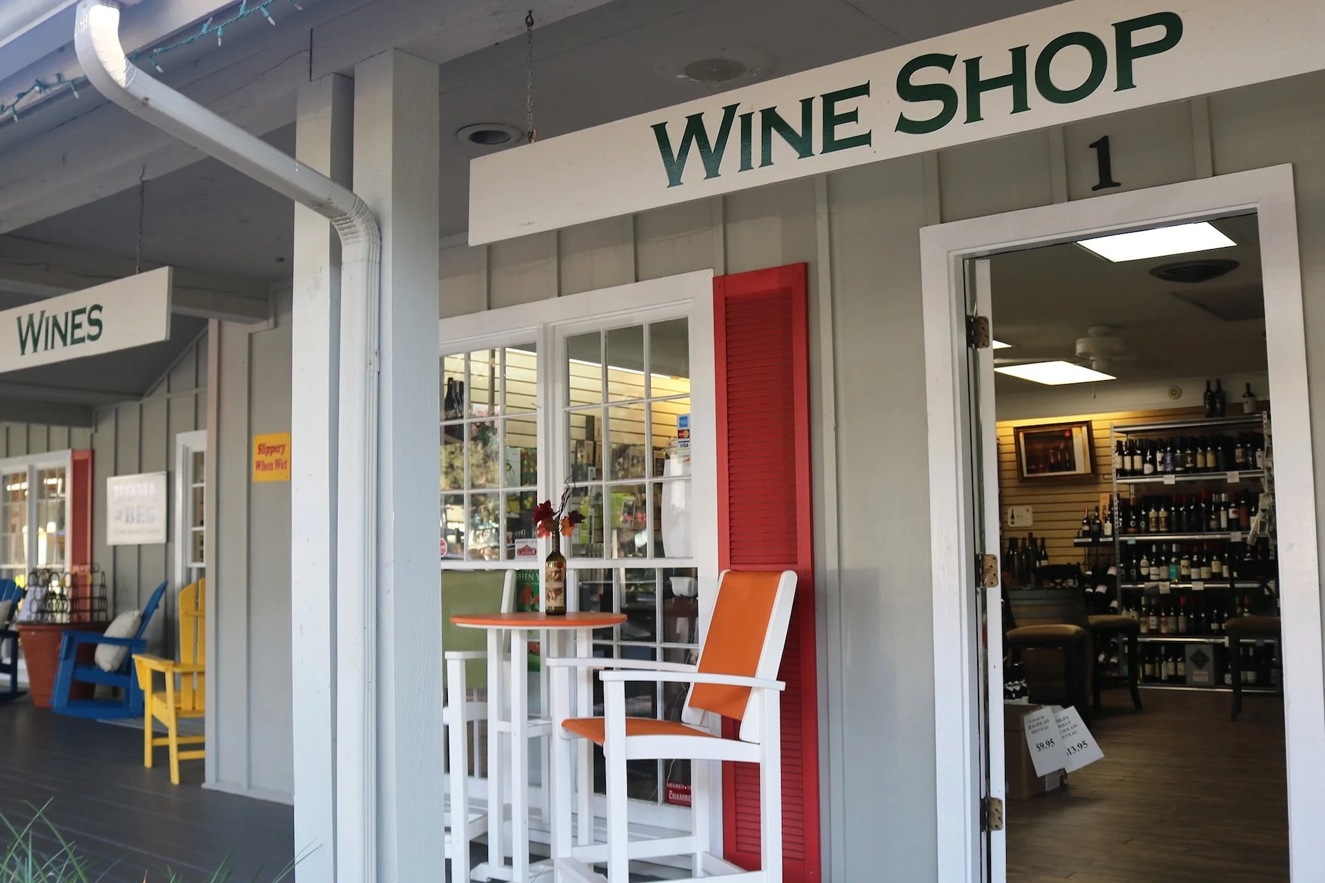 A wine shop with a gray exterior, red shutters, and a sign reading “WINE SHOP” above the entrance. An orange and white table with chairs sits outside, and wine bottles are visible through the shop’s open door and windows.