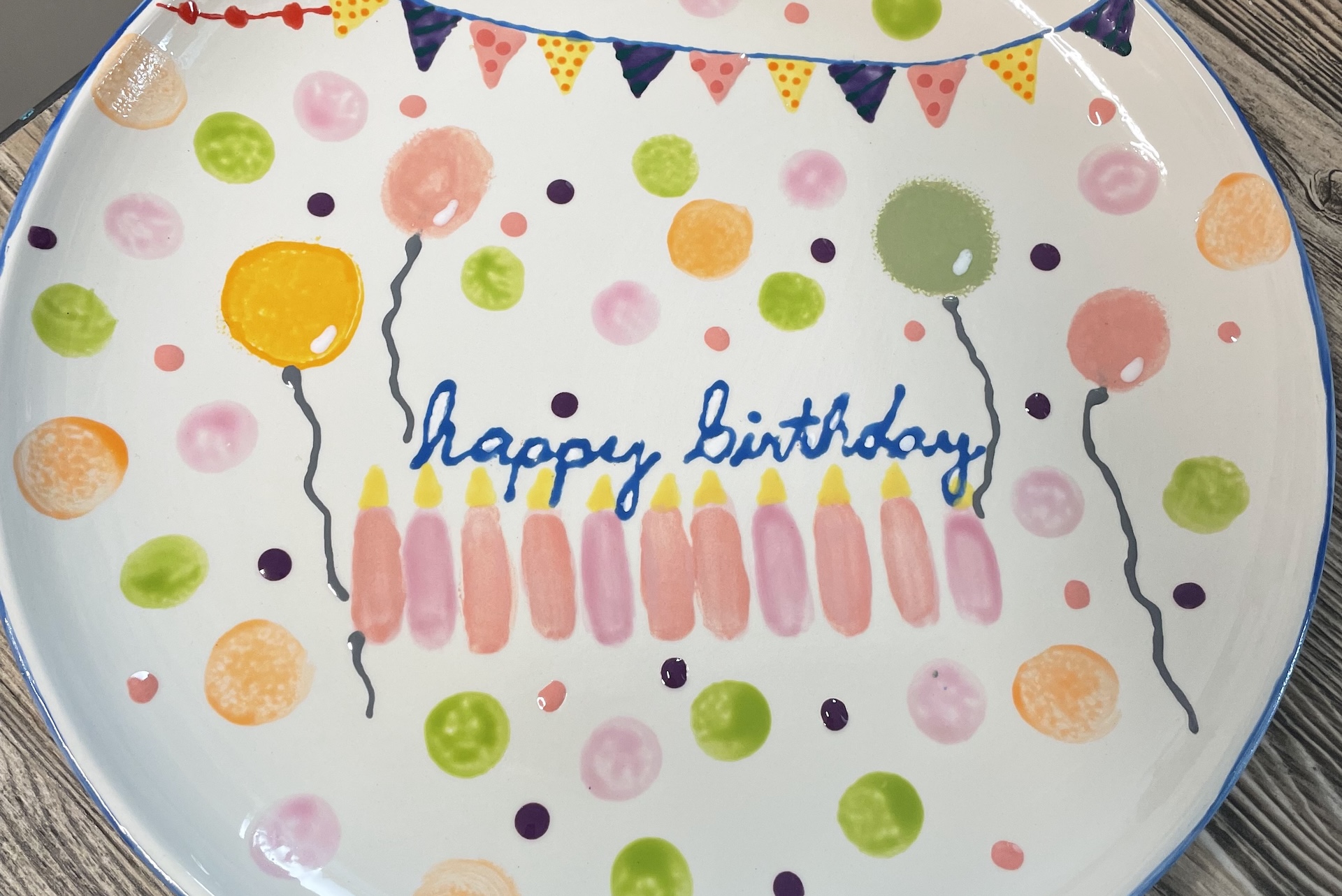 A colorful plate with painted balloons, confetti, a banner, and Happy Birthday written in blue surrounded by ten pink candles. The background is white with multicolored dots.